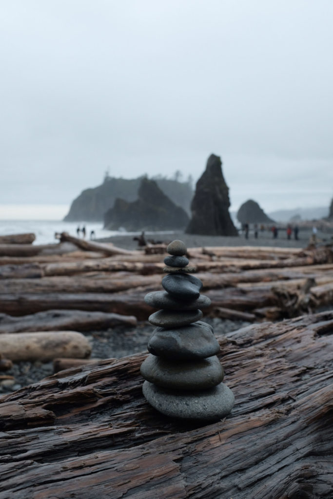 Rock cairns in focus against a backdrop of the rocky, Oregon coast with giant logs.